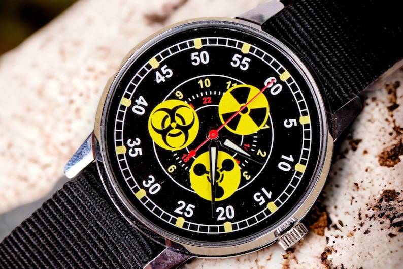 May include: A black wristwatch with a black nylon band. The watch face features yellow hazard symbols and white numerals against a black background. The watch has a silver-colored metal case.