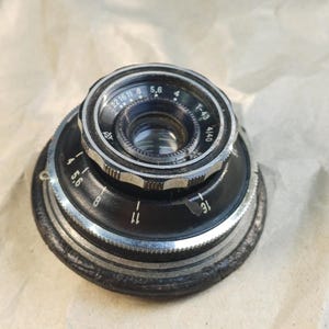 以下が含まれることがあります： 銀色のリングと「22 16 11 8 5,6 4 T-43 4/40 5,6 8 11 16」の刻印がされた黒と銀のカメラレンズ。