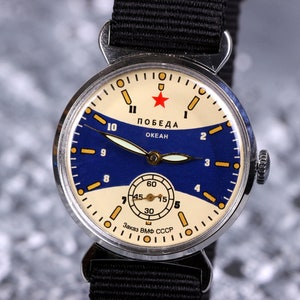 May include: A silver wristwatch with a blue and white face. The watch face has a red star and the words "ПОБЕДА ОКЕАН" in Cyrillic script. The watch has a black nylon strap.
