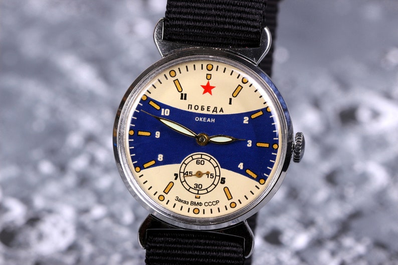 May include: A silver wristwatch with a blue and white face. The watch face has a red star and the words "ПОБЕДА ОКЕАН" in Cyrillic script. The watch has a black nylon strap.
