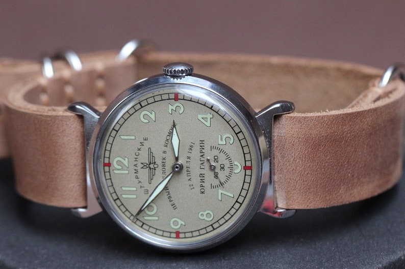 Puede incluir: Un reloj de pulsera plateado con una esfera de color crema y agujas negras. La esfera del reloj tiene marcadores de hora romanos negros y un peque&ntilde;o dial de segundos. La esfera del reloj tambi&eacute;n tiene el texto "ШТУРМАНСКИЕ" y "ПЕРВЫЙ ЧЕЛОВЕК В КОСМОС 12 АПРЕЛЯ 1961 ЮРИЙ ГАГАРИН" en escritura cir&iacute;lica.