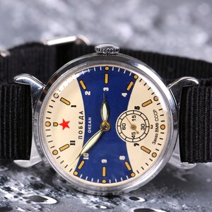 May include: A silver wristwatch with a black nylon strap. The watch face is white with a blue and yellow design. The watch face has the text "ПОБЕДА ОКЕАН Завод ВМФ СССР".