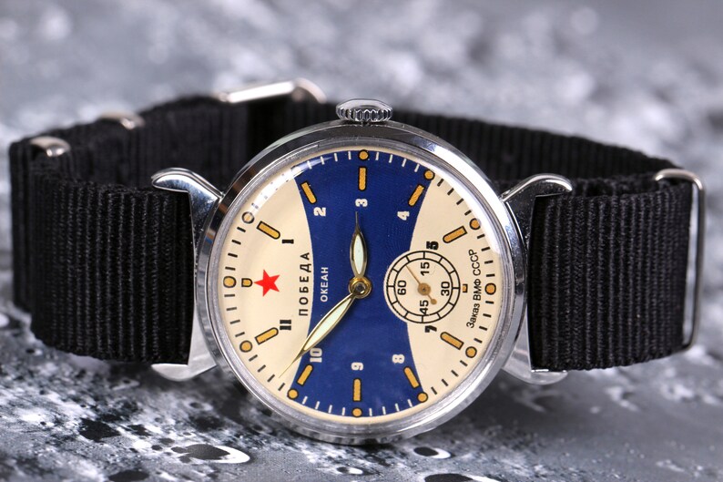 May include: A silver wristwatch with a black nylon strap. The watch face is white with a blue and yellow design. The watch face has the text "ПОБЕДА ОКЕАН Завод ВМФ СССР".