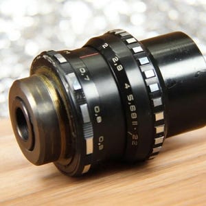 May include: A black camera lens with a silver aperture ring and depth of field markings. The lens has a silver ring with the text "2.8 4 5.6 8 11 22" and "S.68 II" printed on it.