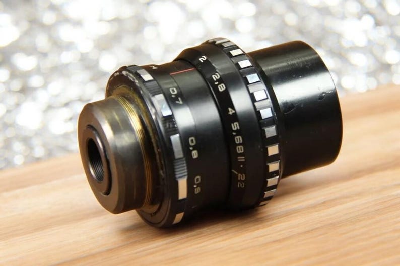 May include: A black camera lens with a silver aperture ring and depth of field markings. The lens has a silver ring with the text "2.8 4 5.6 8 11 22" and "S.68 II" printed on it.