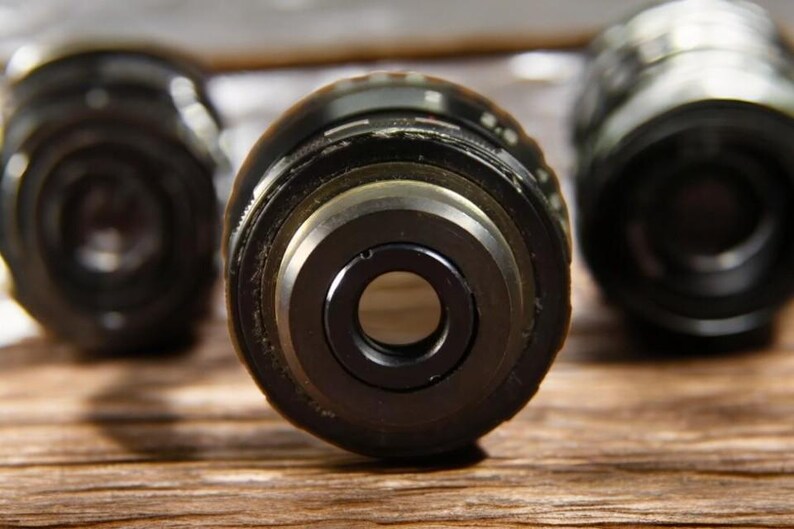 May include: Three black camera lens caps with a silver ring around the opening. The lens caps are on a wooden surface.