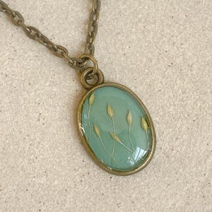Könnte beinhalten: Ovale Anhänger Halskette mit hellgrünem Hintergrund und getrockneten Gräsern. Der Anhänger ist in einem goldfarbenen Metallrahmen gefasst und hängt an einer passenden Kette. Die Halskette wird auf einer strukturierten, neutralfarbenen Oberfläche präsentiert.
