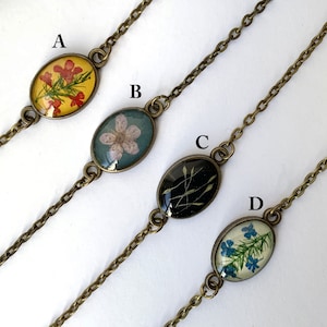 以下が含まれることがあります： 花を押し花にした、楕円形のチャームが付いた繊細なブレスレットが4つ。チャームは黄色、青、白、黒の色合いです。ブレスレットはブロンズ色のチェーンで作られています。