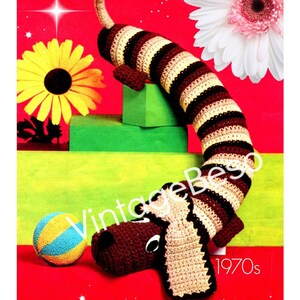 Könnte beinhalten: Gehäkeltes Dackel-Hundespielzeug in braunen, beigen und schwarzen Streifen, mit passendem Kopf und einem kleinen blau-gelben Ball. Das Bild hat eine Retro-Ästhetik, mit rotem Hintergrund und dem Text "1970er Jahre".