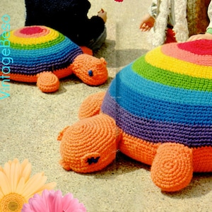 Puede incluir: Dos cojines de suelo en forma de tortuga de crochet arcoíris. Los cojines están hechos con una variedad de colores, incluyendo rojo, naranja, amarillo, verde, azul, índigo y violeta. Los cojines están sentados en una superficie arenosa. La imagen incluye el texto "vintagebeso 1970s".