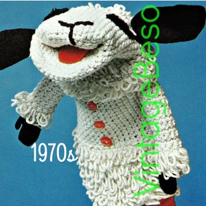May include: A white crocheted sheep puppet with black ears and a red tongue. The puppet has three red buttons on its chest and the text "1970s" is visible in the lower left corner of the image.