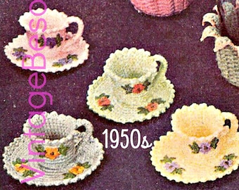 May include: A set of four crocheted teacups and saucers with floral designs. The teacups are in shades of pink, white, and yellow. The saucers have a scalloped edge and are decorated with small flowers. The text "1950s" is visible in the image.