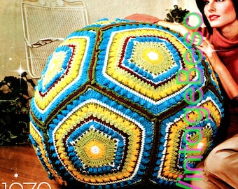May include: A large, crocheted, multi-colored, hexagonal-shaped floor pillow. The pillow is made with yellow, blue, green, white, and brown yarn. The pillow is sitting on a brown floor.