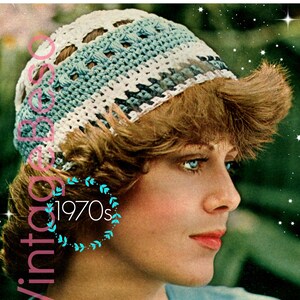 Op de afbeelding: Een vrouw draagt een witte en blauwe gehaakte hoed met een ontwerp uit de jaren 1970. De hoed heeft een kantpatroon en wordt gedragen over haar bruine haar.