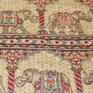 May include: Beige fabric with a repeating pattern of elephants and floral designs. The elephants are depicted in a traditional Indian style with intricate details and are carrying ornate saddles. The fabric has a subtle sheen and a textured surface.