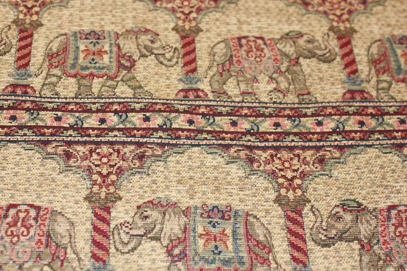 May include: Beige fabric with a repeating pattern of elephants and floral designs. The elephants are depicted in a traditional Indian style with intricate details and are carrying ornate saddles. The fabric has a subtle sheen and a textured surface.