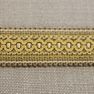 May include: Close-up of decorative trim with a repeating pattern. The trim features a central band of olive green loops, bordered by thinner bands of beige and gold. The background is a textured, light gray fabric.