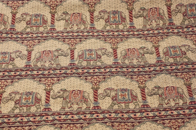 May include: Beige fabric with a repeating pattern of elephants walking under arches. The elephants are decorated with colourful floral designs and are outlined in brown. The arches are made of red and white stripes.