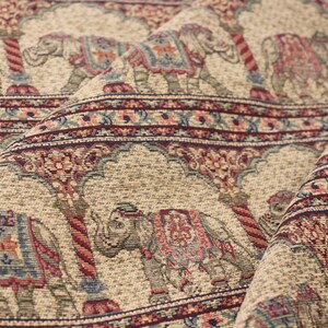 May include: Beige fabric with a repeating pattern of elephants in a colourful floral design. The elephants are carrying ornate saddles and are walking under a decorative archway.