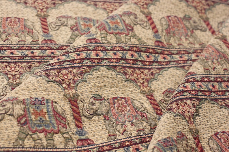 May include: Beige fabric with a repeating pattern of elephants in a colourful floral design. The elephants are carrying ornate saddles and are walking under a decorative archway.