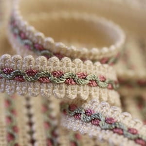 May include: Close-up of a cream-colored decorative trim with a scalloped edge. The trim features a woven design with alternating sections of pink and green threads, creating a delicate and textured appearance. The trim is likely used for sewing or crafting.