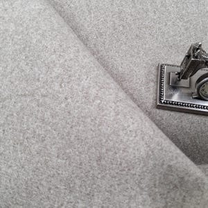 May include: A silver-colored miniature sewing machine with a clock face, resting on a textured gray fabric. The sewing machine is detailed with intricate designs, suggesting a vintage aesthetic. The fabric has a soft, felt-like appearance.