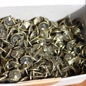 May include: A box full of gold-coloured decorative tacks. The tacks have a round head with a decorative design and a pointed tip.