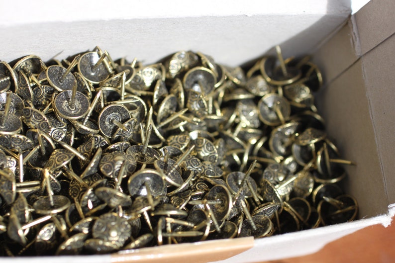 May include: A box full of gold-coloured decorative tacks. The tacks have a round head with a decorative design and a pointed tip.