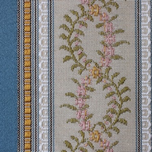 May include: Close-up of a woven fabric with a repeating vertical stripe pattern. The design features alternating stripes of blue, beige, and gold. Floral patterns in pink and green adorn the beige stripes, creating a textured, decorative textile.