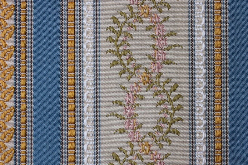 May include: Close-up of a woven fabric with a repeating vertical stripe pattern. The design features alternating stripes of blue, beige, and gold. Floral patterns in pink and green adorn the beige stripes, creating a textured, decorative textile.