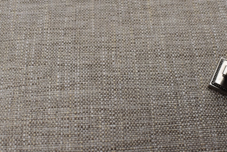 May include: A close-up of a beige textured fabric with a silver metal decorative piece in the bottom right corner.