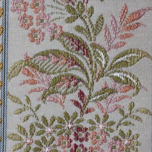 May include: Close-up of a woven fabric with a floral pattern. The design features pink, green, and peach-colored flowers and leaves on a light gray background. The edges have a blue border and gold leaf-like accents.