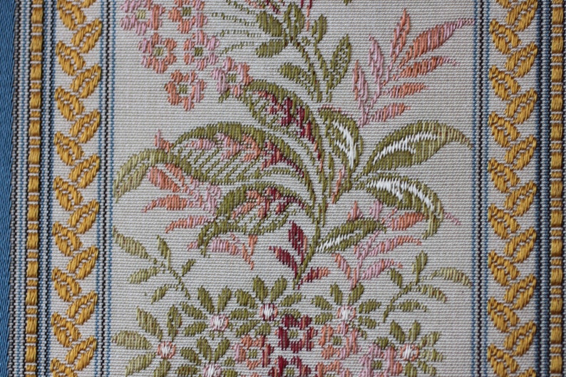 May include: Close-up of a woven fabric with a floral pattern. The design features pink, green, and peach-colored flowers and leaves on a light gray background. The edges have a blue border and gold leaf-like accents.