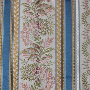 May include: Close-up of a patterned fabric with vertical stripes. The design features alternating stripes of light blue, beige, and floral patterns in pink, green, and gold. The fabric has a textured appearance, suitable for upholstery or decorative use.