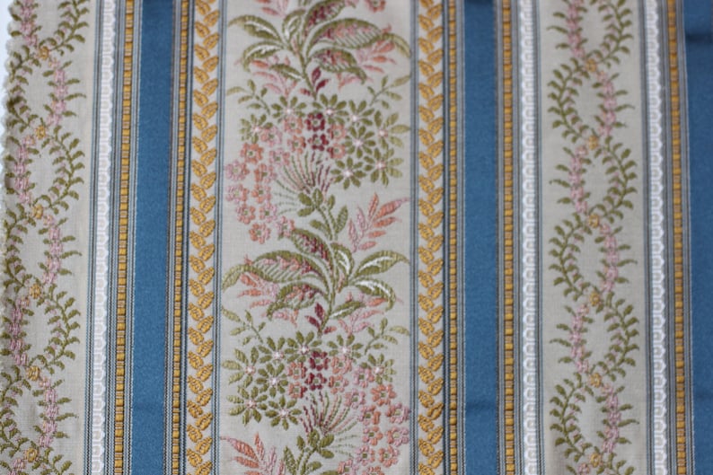 May include: Close-up of a patterned fabric with vertical stripes. The design features alternating stripes of light blue, beige, and floral patterns in pink, green, and gold. The fabric has a textured appearance, suitable for upholstery or decorative use.