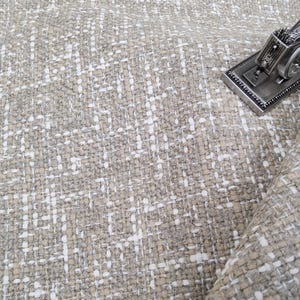 May include: A textured fabric in shades of beige and white, with a small decorative sewing machine figurine. The fabric has a woven appearance, with a grid-like pattern. The sewing machine is silver-colored.