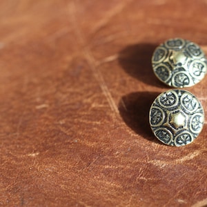 May include: Two gold-toned metal buttons with a decorative black design. The buttons have a raised centre and a circular shape.