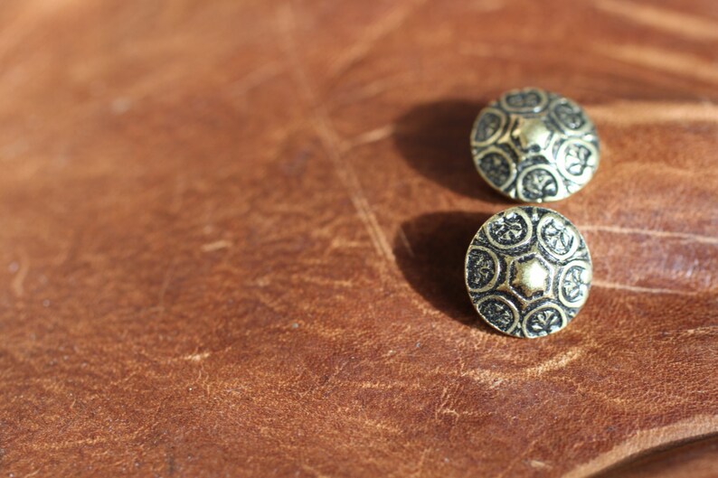 May include: Two gold-toned metal buttons with a decorative black design. The buttons have a raised centre and a circular shape.