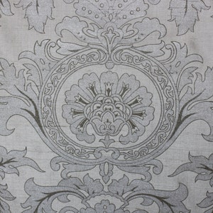 May include: A silver and gray fabric with a repeating pattern of stylized floral designs. The fabric has a subtle sheen and a textured surface.