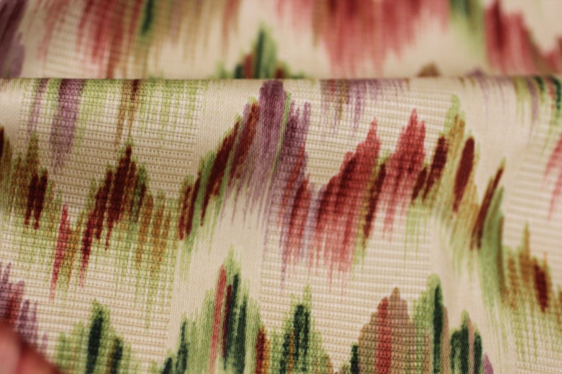 Op de afbeelding: Close-up van een cr&egrave;mekleurige stof met een geweven textuur. De stof heeft een abstract patroon in rood, groen en paarse tinten.