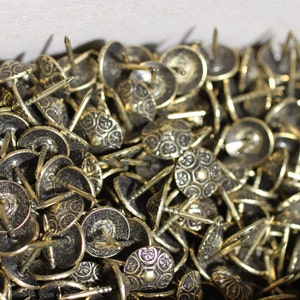 May include: A close-up image of a large pile of decorative brass tacks with ornate designs. The tacks have a shiny, metallic finish and are ideal for adding a touch of elegance to furniture, crafts, and other projects.