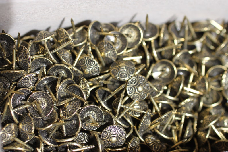 May include: A close-up image of a large pile of decorative brass tacks with ornate designs. The tacks have a shiny, metallic finish and are ideal for adding a touch of elegance to furniture, crafts, and other projects.