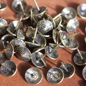 May include: A close-up of a pile of decorative brass tacks with a detailed, ornate design. The tacks have a dark, aged finish and are perfect for adding a touch of vintage charm to furniture, crafts, and home decor.