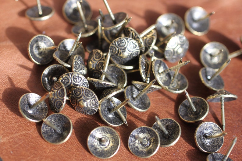 May include: A close-up of a pile of decorative brass tacks with a detailed, ornate design. The tacks have a dark, aged finish and are perfect for adding a touch of vintage charm to furniture, crafts, and home decor.