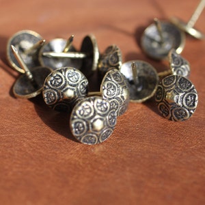May include: A close-up of a group of decorative metal tacks with ornate designs. The tacks are gold-toned and have a pointed end for hammering into surfaces.