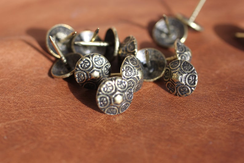 May include: A close-up of a group of decorative metal tacks with ornate designs. The tacks are gold-toned and have a pointed end for hammering into surfaces.