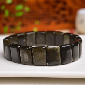 May include: A stretch bracelet composed of rectangular, polished black and gold obsidian beads. The beads feature a smooth, reflective surface with subtle metallic golden sheen inclusions visible within the dark, opaque stone segments.