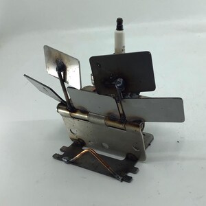 May include: A handcrafted metal sculpture featuring a spark plug and hinged metal plates. The sculpture has a unique, industrial aesthetic with a mix of silver and dark metal tones. The design incorporates rectangular and square shapes, creating a geometric composition.