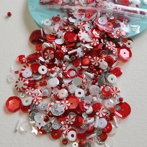 May include: A mix of red, white, and silver craft supplies, including sequins, beads, and star-shaped embellishments. The assortment features various shapes and sizes, with a focus on circular and peppermint candy designs. Some items are spilling from a clear plastic bag.