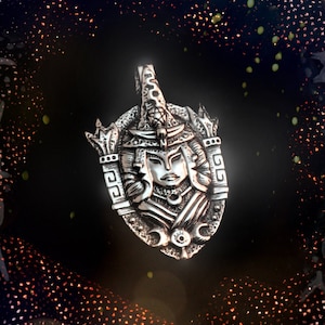May include: A silver pendant with a detailed design of a woman's face with three faces, surrounded by a decorative frame. The pendant is on a black background with a glittery effect.